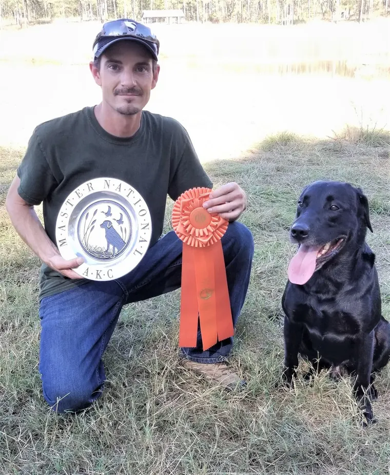 Richard Gebhart with an AKC Master National plate, an orange ribbon and a black Labrador